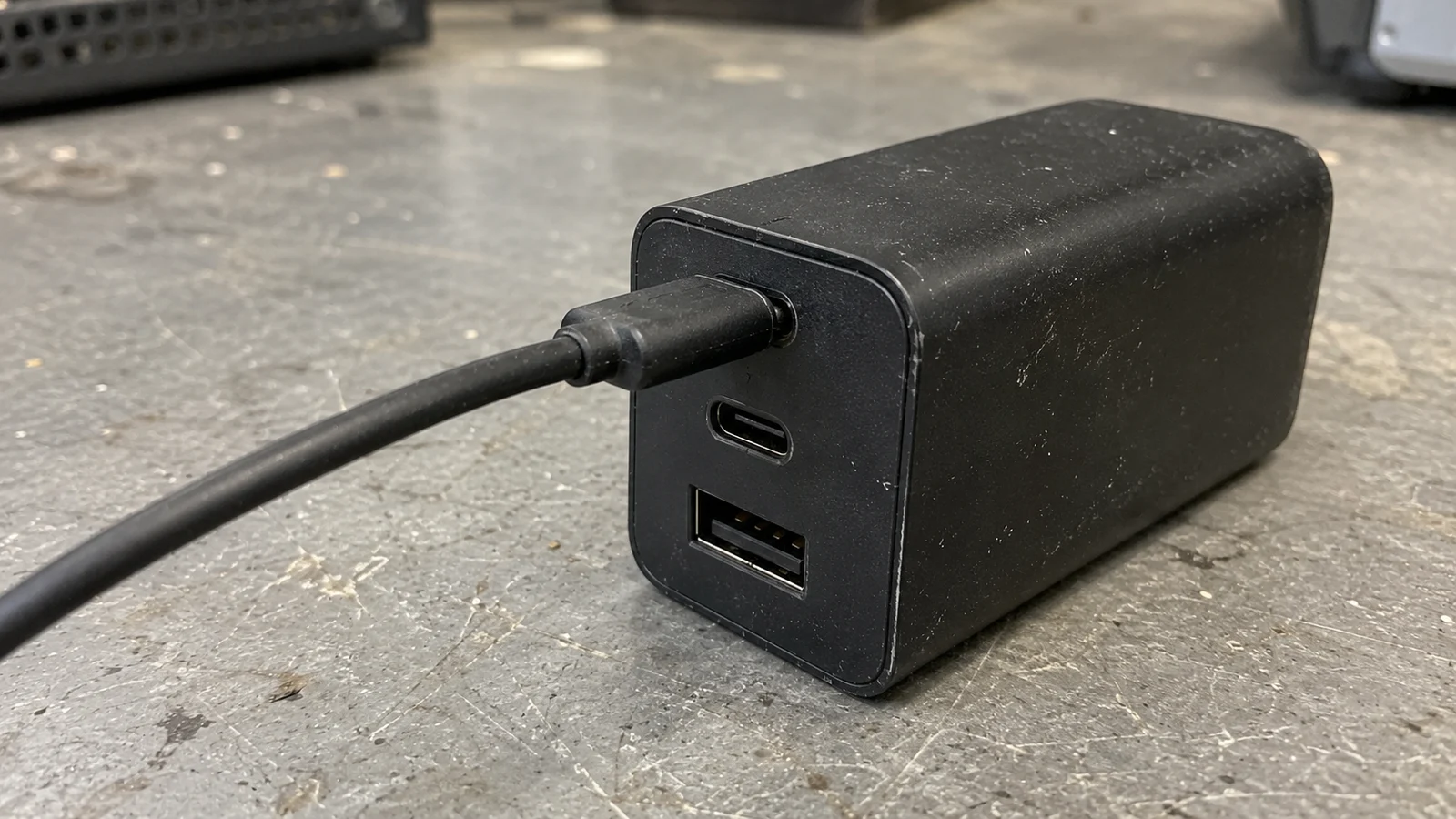 A dark three-port charger with a cable connected to the upper USB-C socket on a worn grey bench